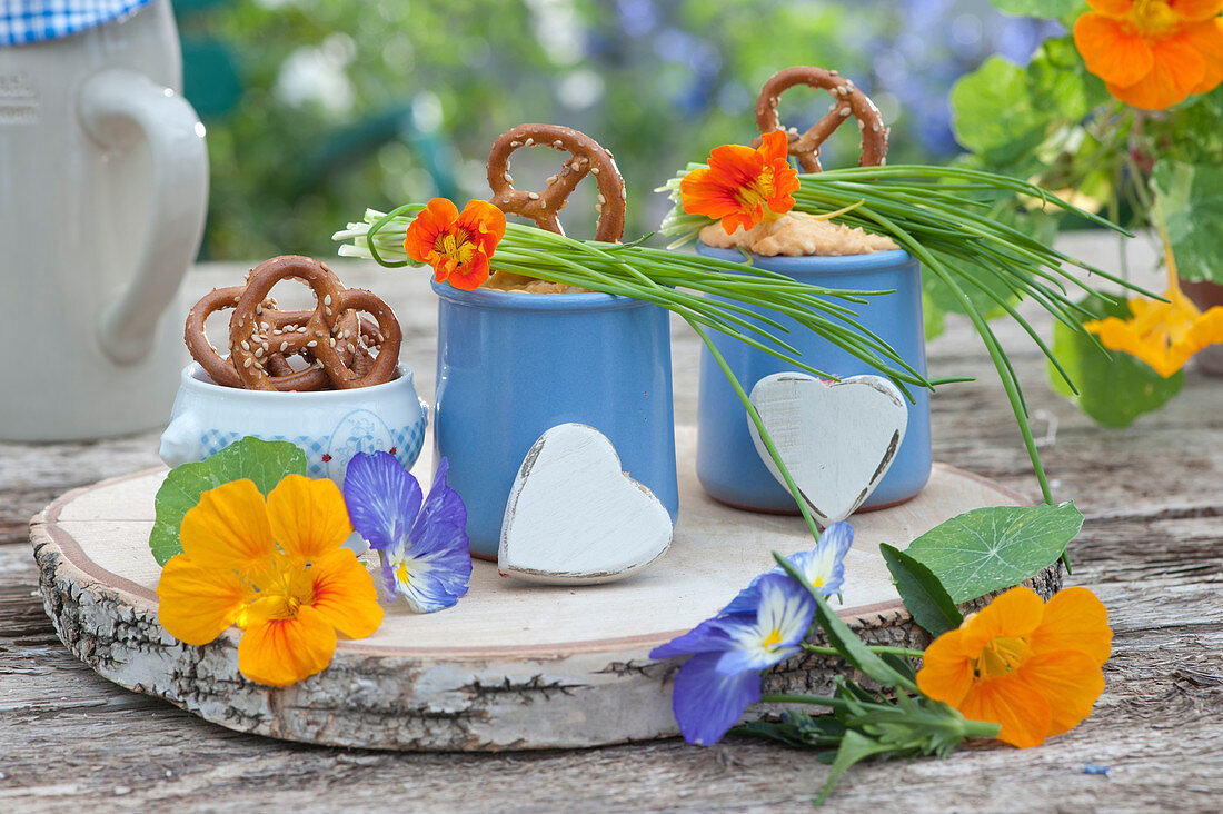Mug with obatzder, pretzels, chives, flowers of nasturtium and horned violets on a wooden board made of birch wood, wooden hearts as decoration
