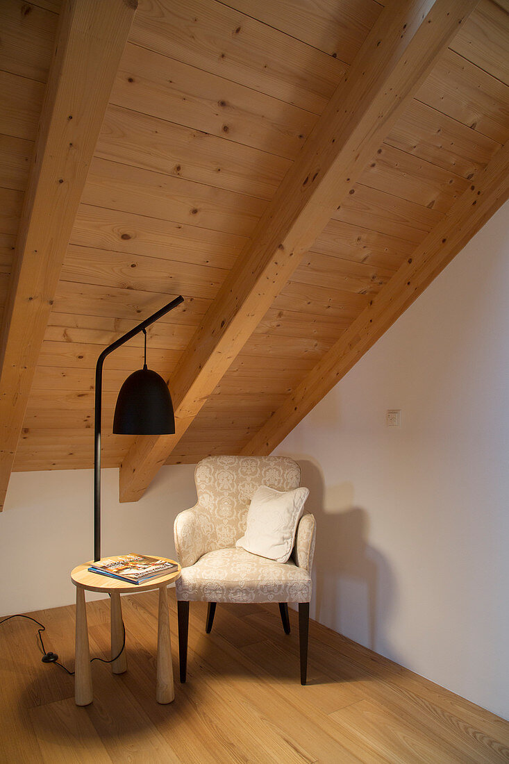 Armchair, standard lamp and side table below wood-clad sloping ceiling
