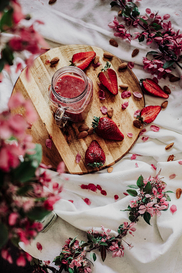 Glaskrug mit Erdbeersmoothie, umgeben von Erdbeeren und Blüten