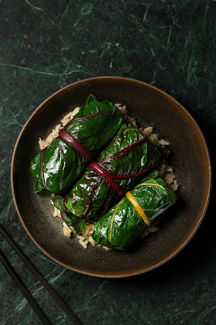 Japanese inspired rainbow chard rolls, filled with mushrooms and finished in a miso broth. They are served on brown rice in a brown stoneware bowl, with chopsticks alongside.
