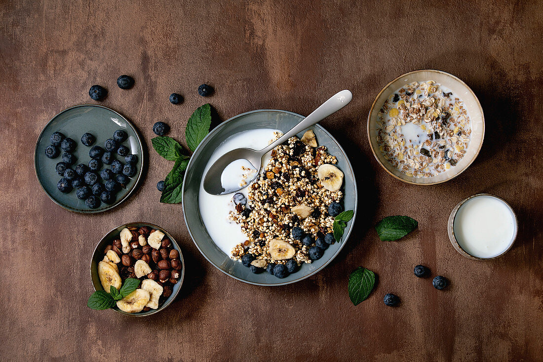 Homemade crunchy puffed millet grain granola with dried fruits and nuts in ceramic bowl, with yogurt, mint and ingredients above. Brown texture background. Flat lay, space. Healthy food breakfast