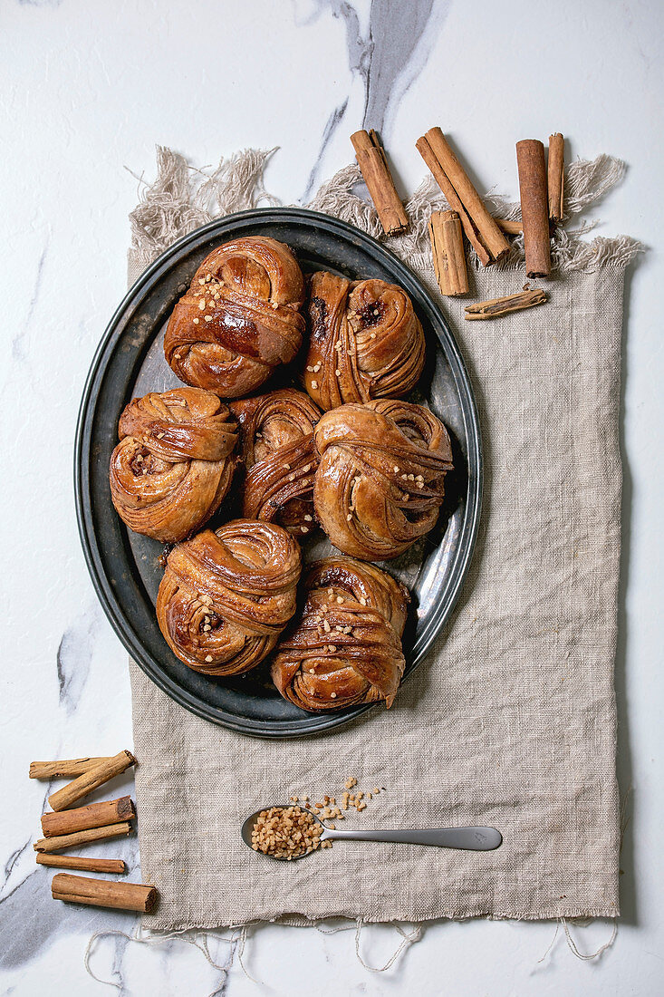 Traditional Swedish cinnamon sweet buns Kanelbulle on vintage tray, cinnamon sticks on linen cloth over white marble background. Flat lay, space.