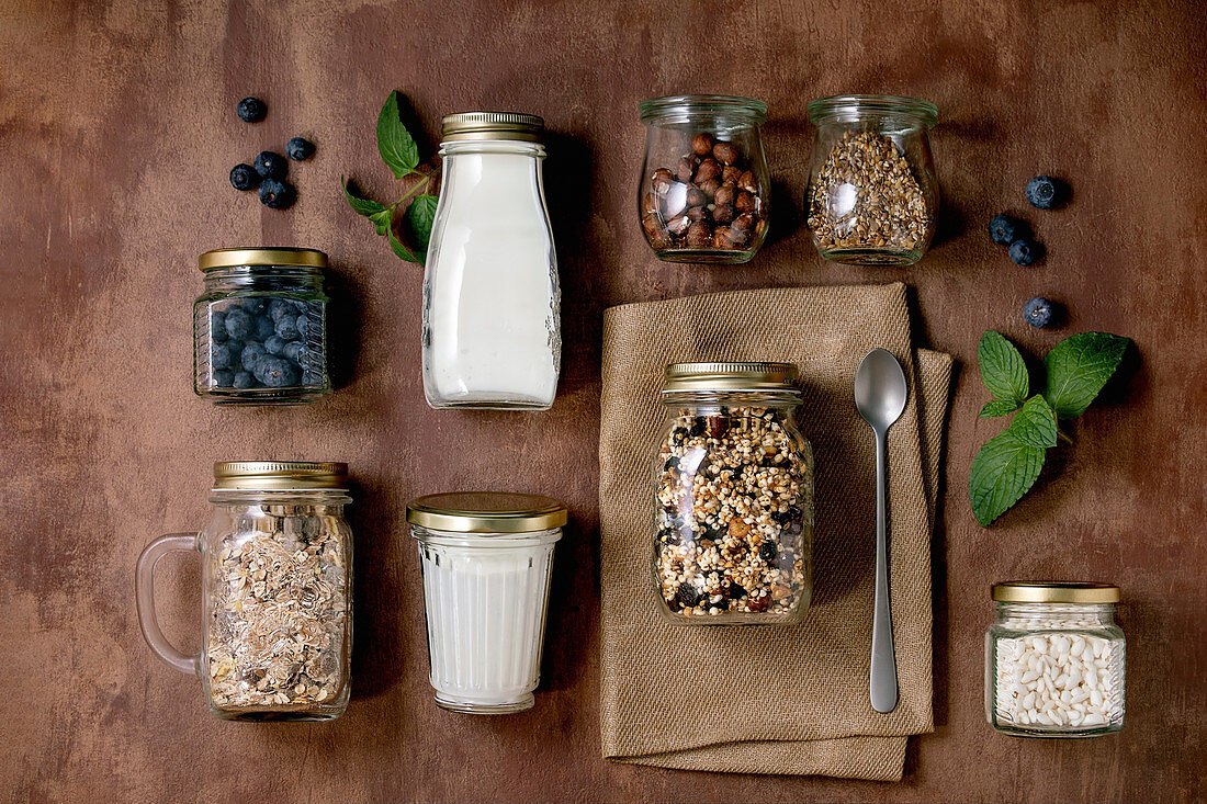 Homemade crunchy puffed millet grain granola with dried fruits and nuts, yogurt, milk, muesli in different glass jars, mint. Brown background. Flat lay, space. Healthy food eco friendly breakfast