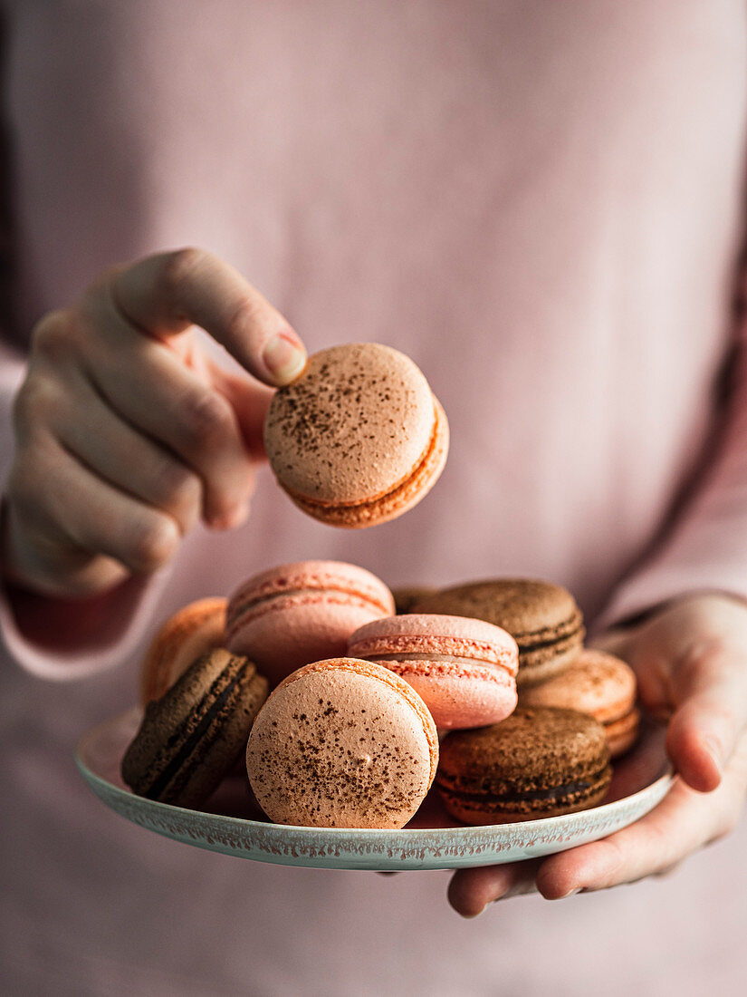 Heap of french macarons on pink plate in hands of woman dressed in pink clothes. Vertical. Copy space