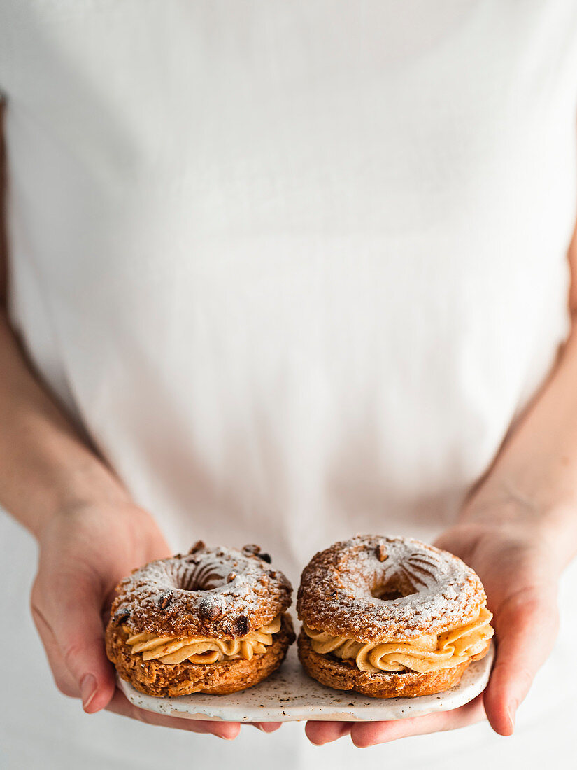 Woman in white t-shirt holds plate with two Choux Paris Brest pastry with hazelnut