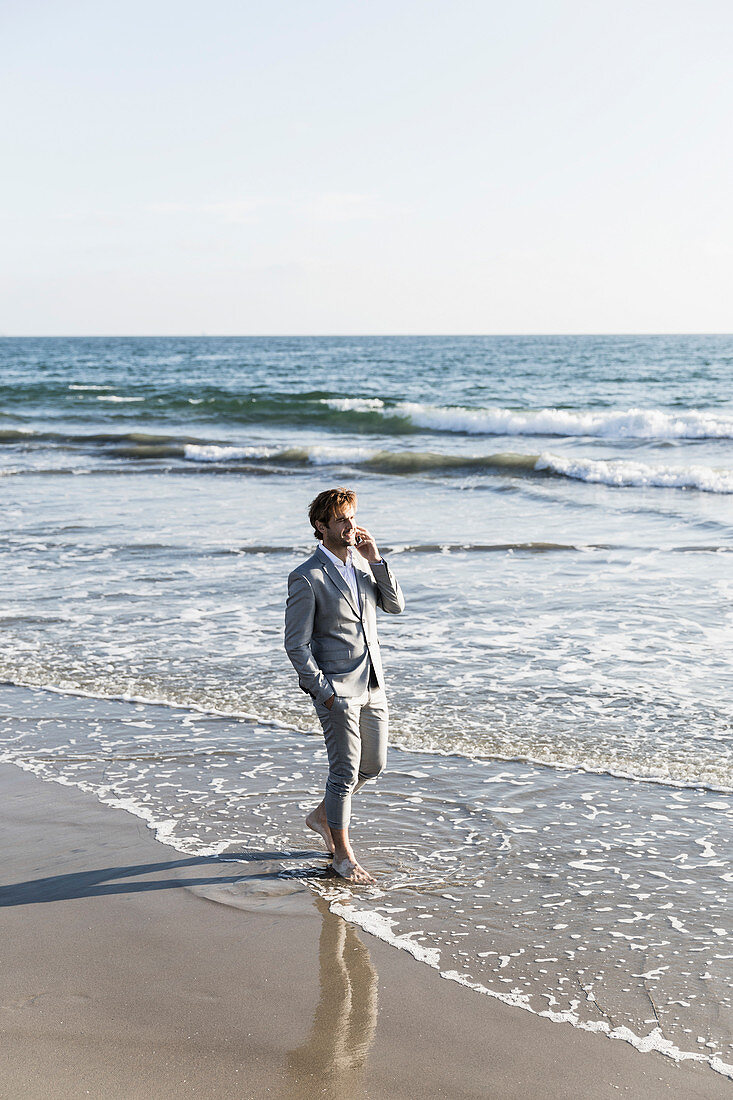 Barefoot businessman talking on smart phone in sunny ocean surf, Los Angeles, California