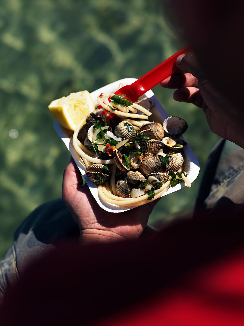 A boy eating spaghetti with clams from a paper dish by the sea