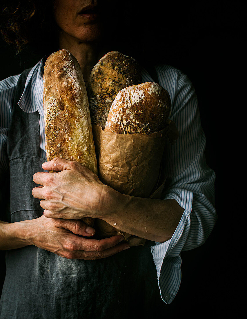 Cropped unrecognizable woman in apron holding ciabatta bread while standing on dark background