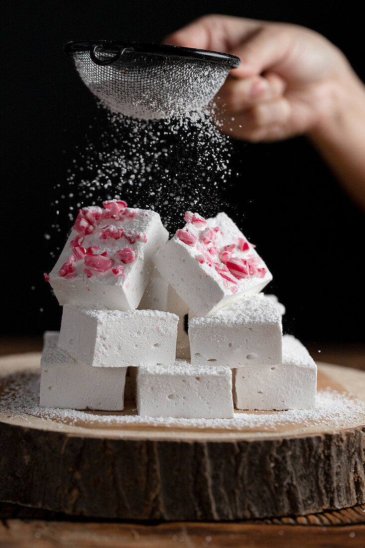 Crop person with sieve sprinkling sugar powder over pieces of marshmallow on wooden board