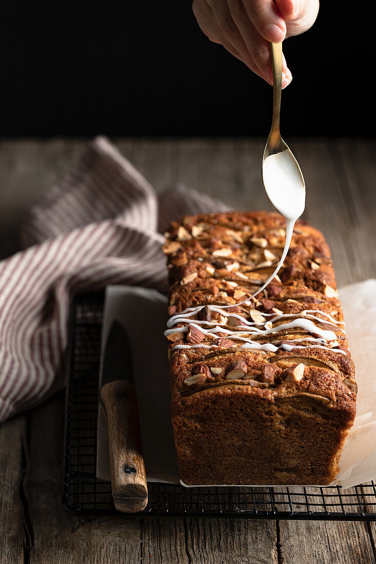 Pouring white sugar icing over homemade banana bread with nuts placed on metal grid on wooden table