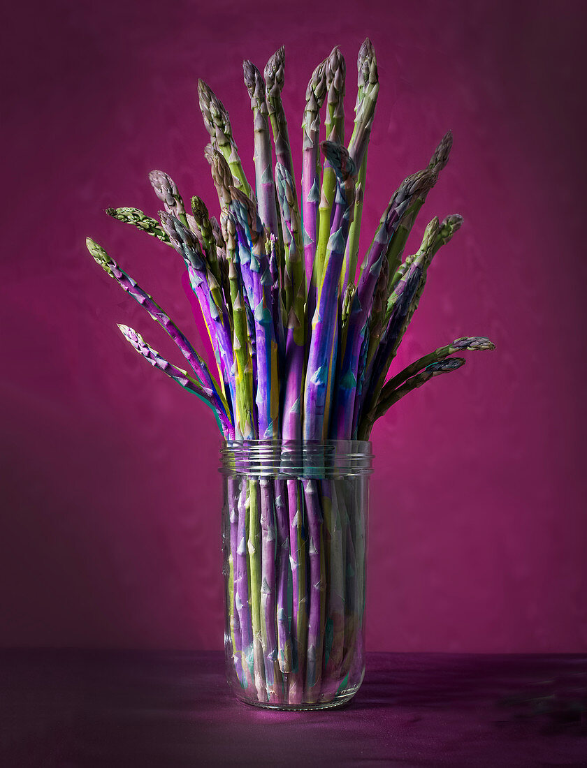Fresh asparagus spears in a glass against a red-violet background