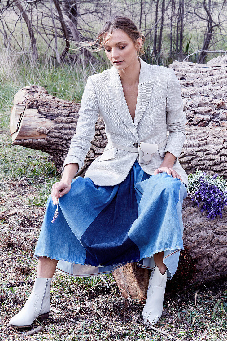 A blonde woman wearing a white blazer and a denim skirt sitting on a fallen tree