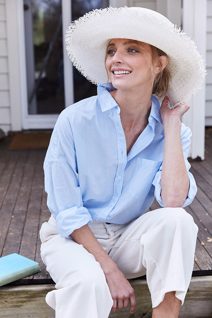 Blonde Frau mit Hut in hellblauer Hemdbluse und weißer Hose sitzt auf der Terrasse