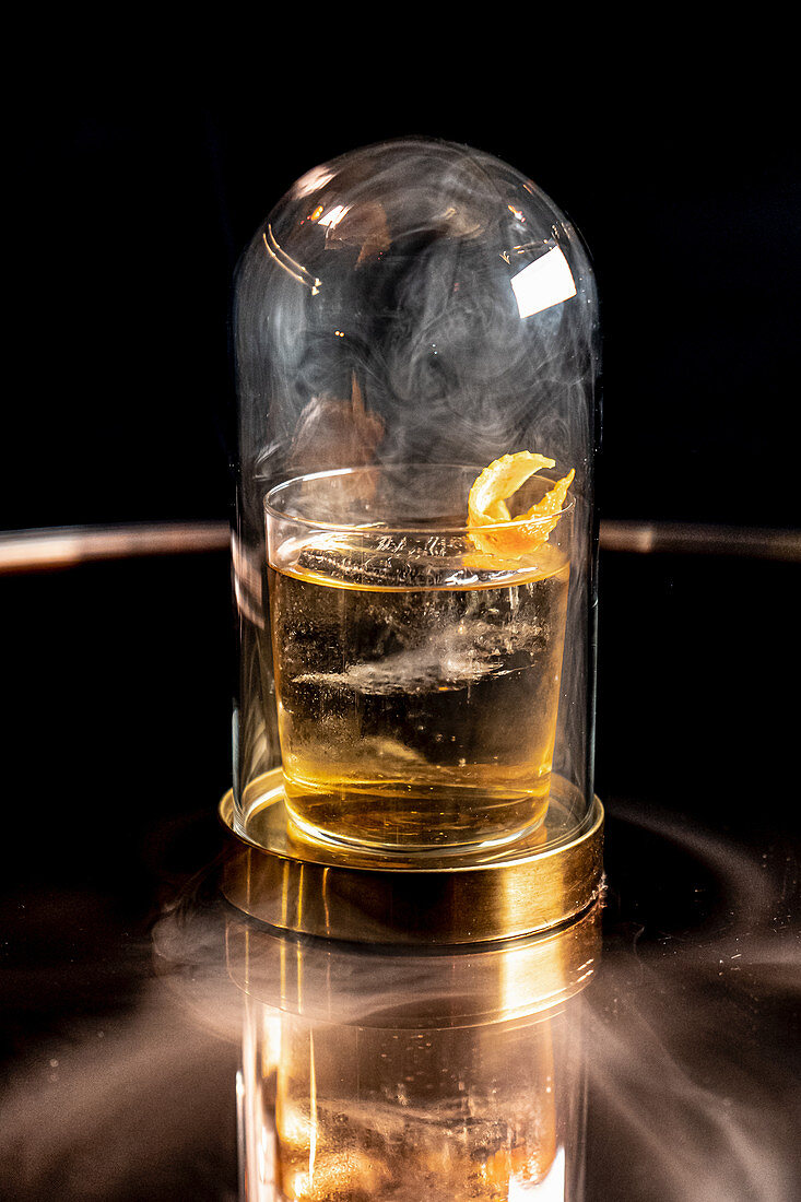 Glass of alcohol drink placed under dome with smoke on reflective table in bar