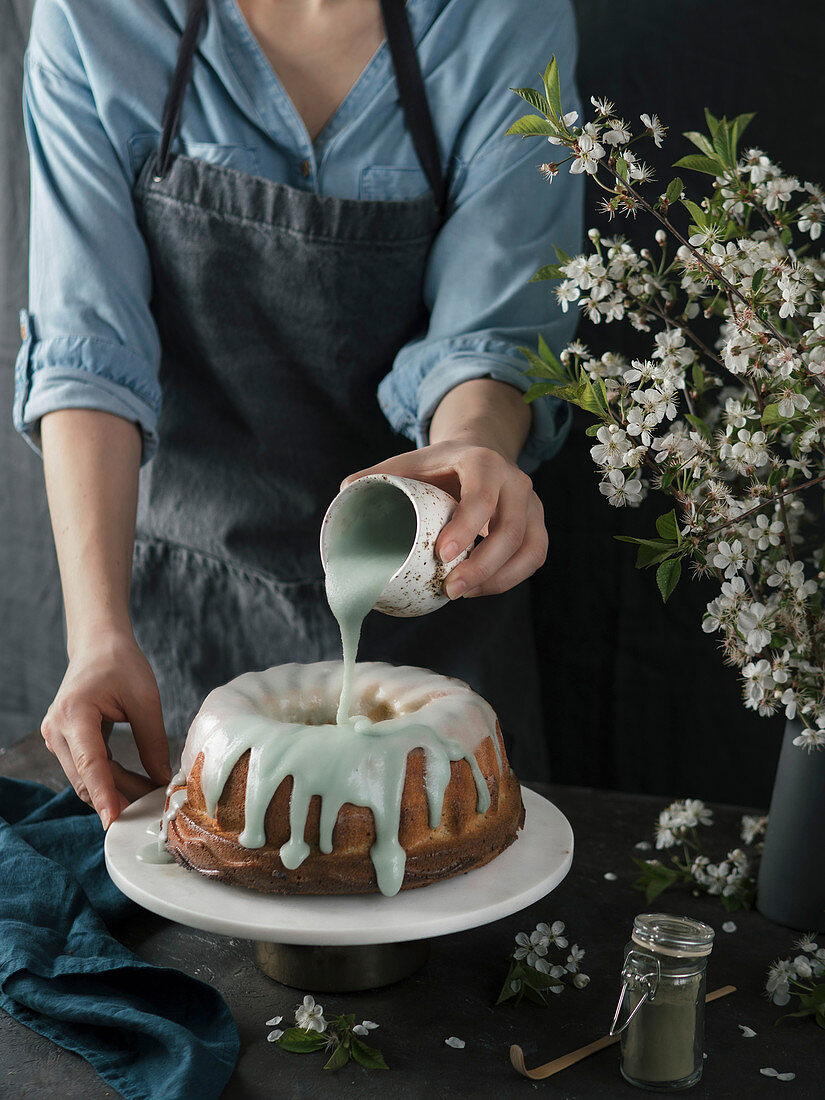 Young woman in blue shirt and gray apron pouring green matcha glaze on bundt cake