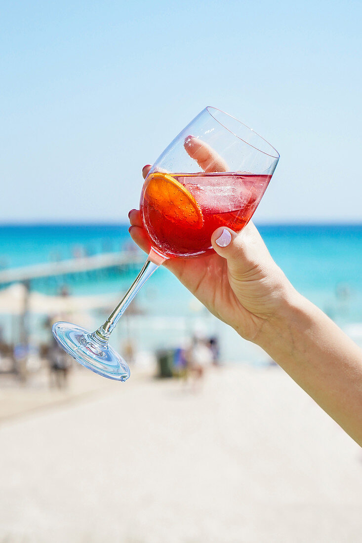 Hand hält Weinglas mit Zitruscocktail am Strand