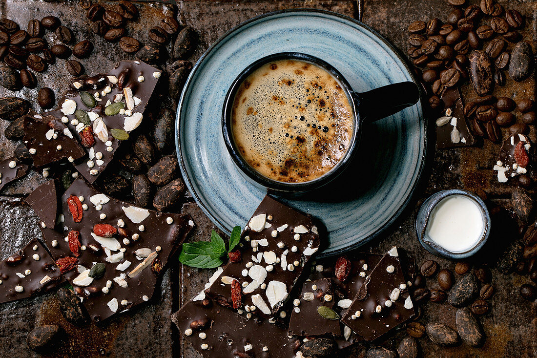Tasse Kaffee inmitten von dunkler Bruchschokolade mit Saaten und Gojibeeren