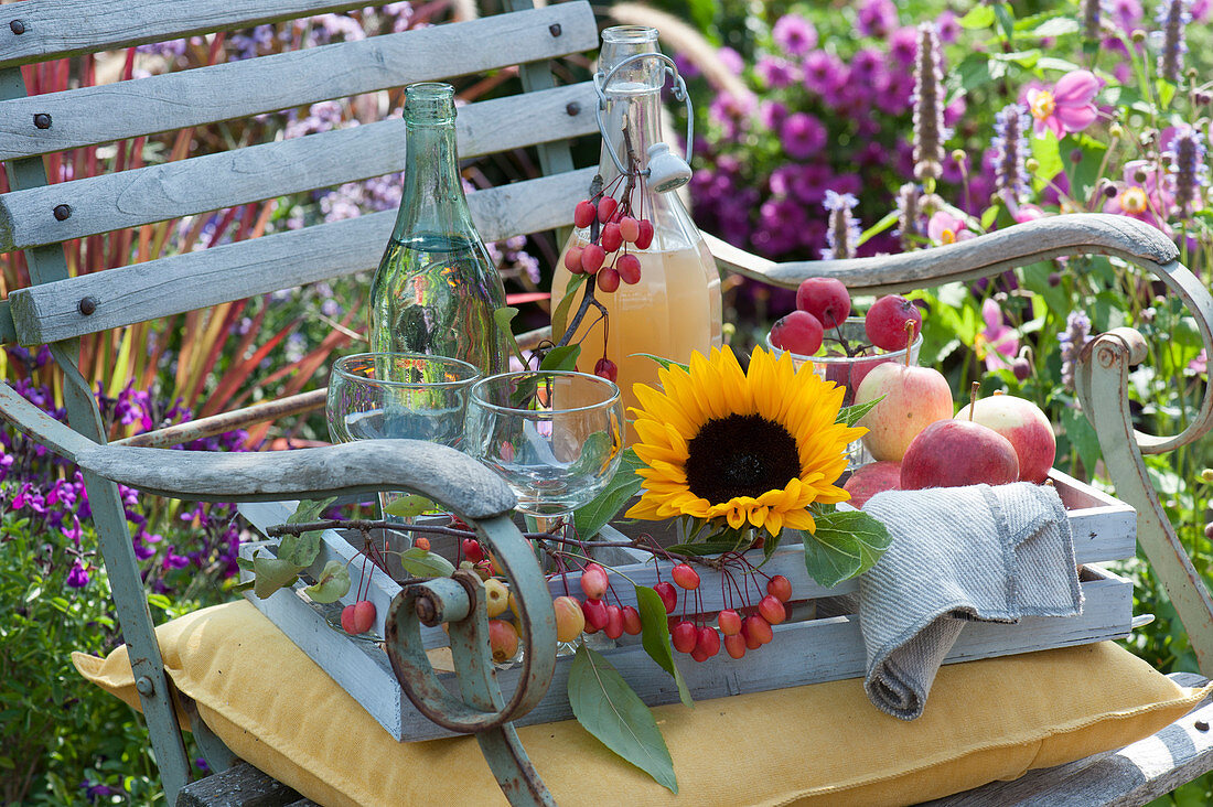 Holzkiste mit Sonnenblume, Äpfeln, Zieräpfeln, Flaschen mit Apfelsaft und Wasser, Gläser