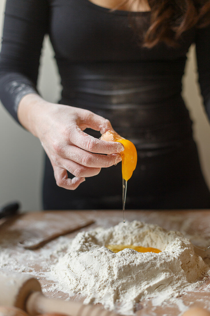 Faceless female in black leather dress breaking raw eggs into flour mass during preparation of pasta dough in kitchen at home in daytime