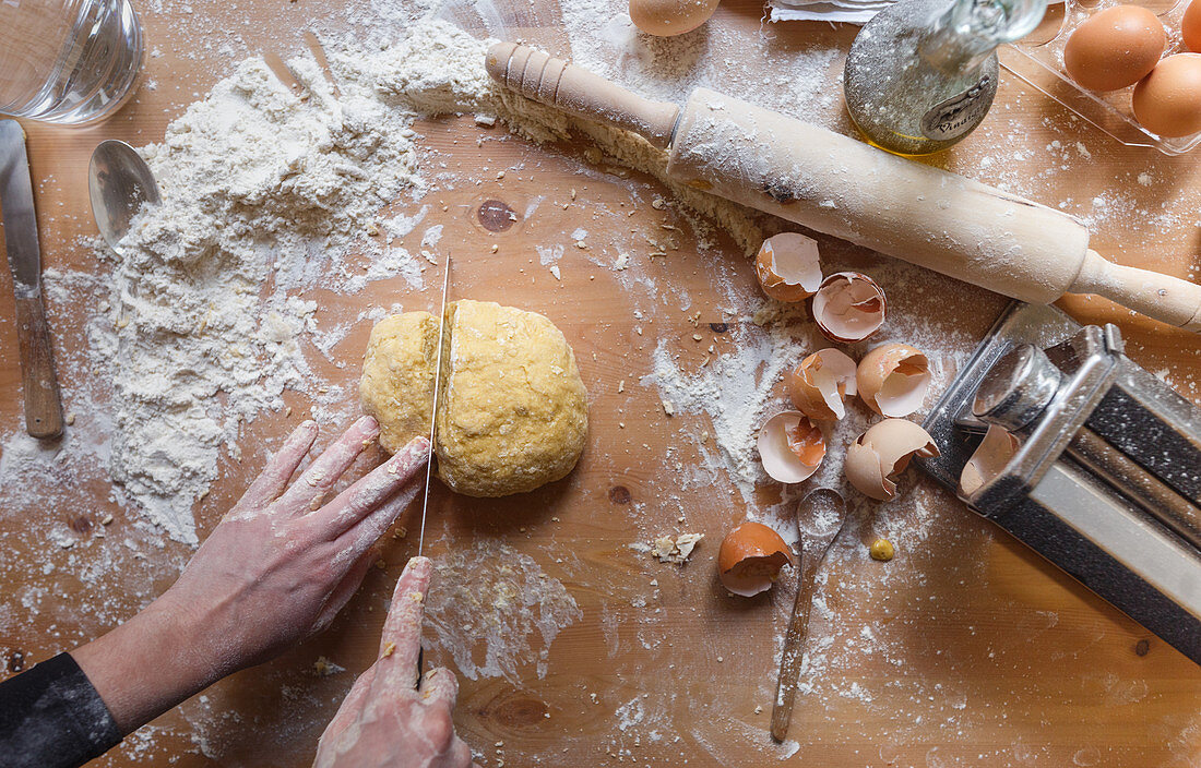 Unrecognizable person cutting dough by long steel knife on table with flour near rolling pin and eggshell while preparing pastry in home kitchen