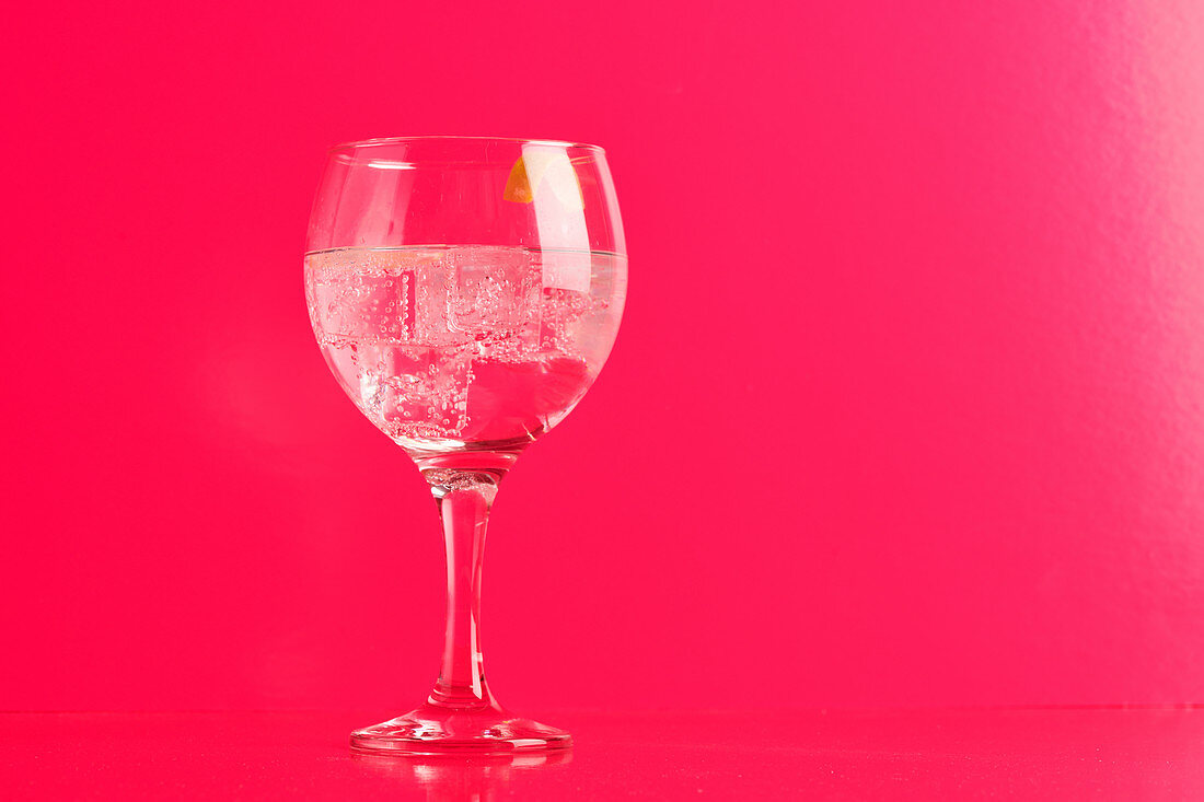 Transparent glass of tasty gin tonic cocktail with ice with lemon twist peel on table in pink background