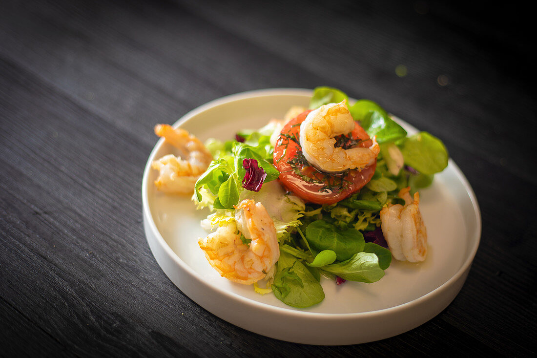 Healthy spinach salad with shrimps and tomato served on withe ceramic plate on black table