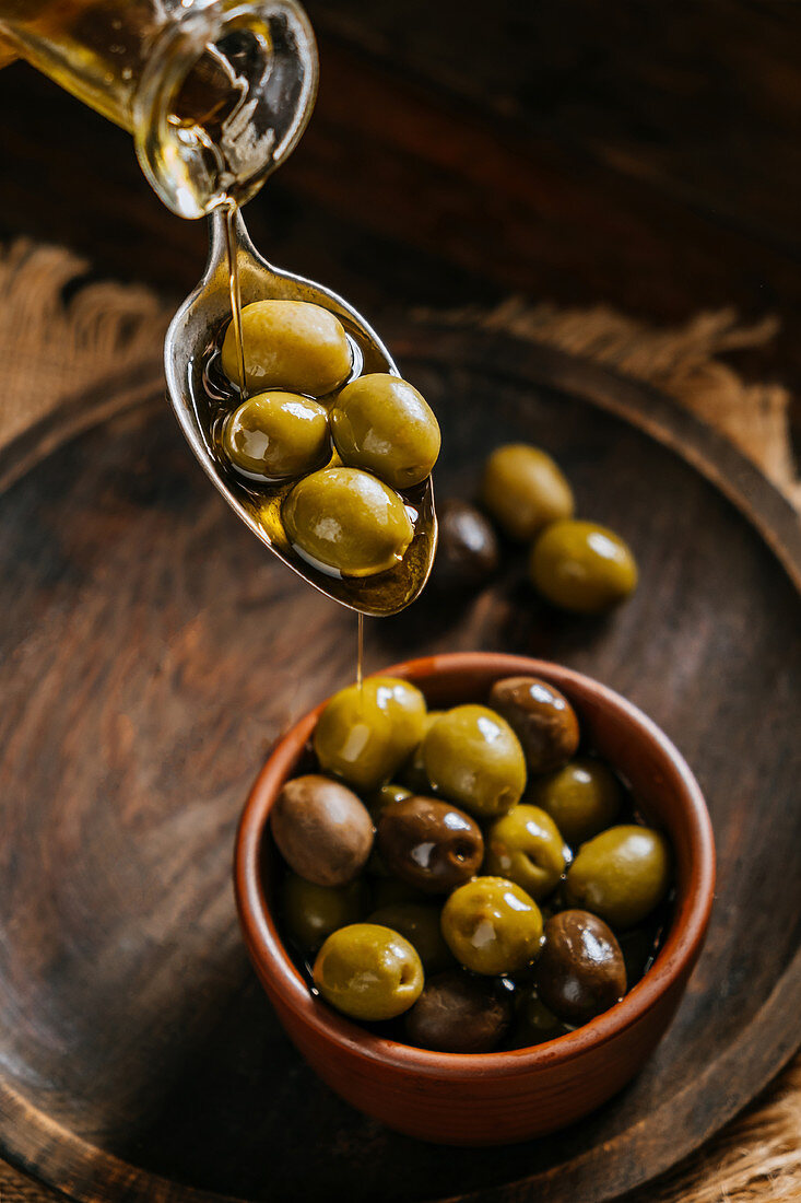 Pouring oil in spoon and ceramic bowl with olives placed on wooden table in kitchen
