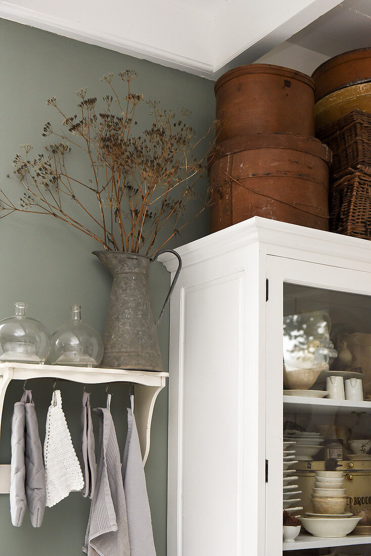 Storage boxes on a white cupboard next to a zinc jug of a dried flowers on a shelf