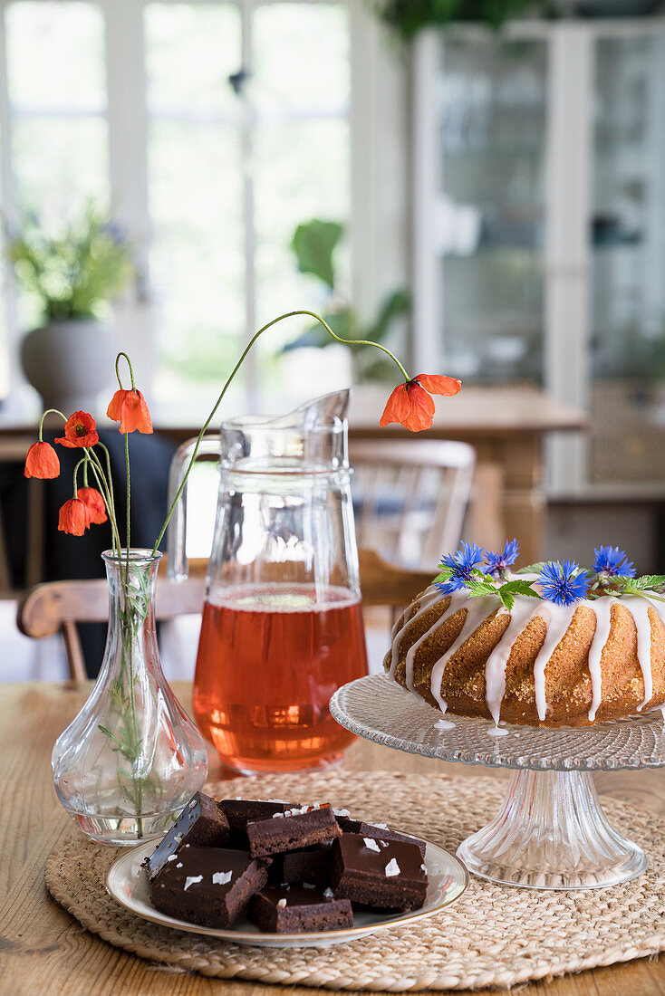 A Bundt cake with icing and flowers, chocolate and a drink on table