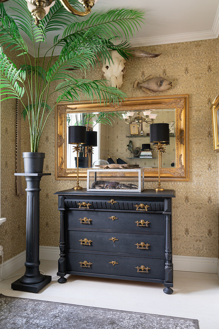 An indoor palm tree on a stela, an antique chest of drawers and a gold-framed mirror on a papered wall