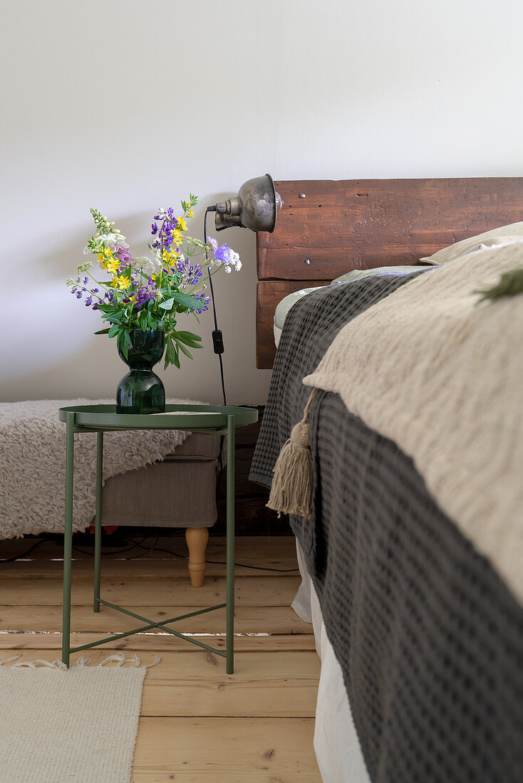 A bouquet of flowers on a bedside table next to a bed with headboard made of recycled wood