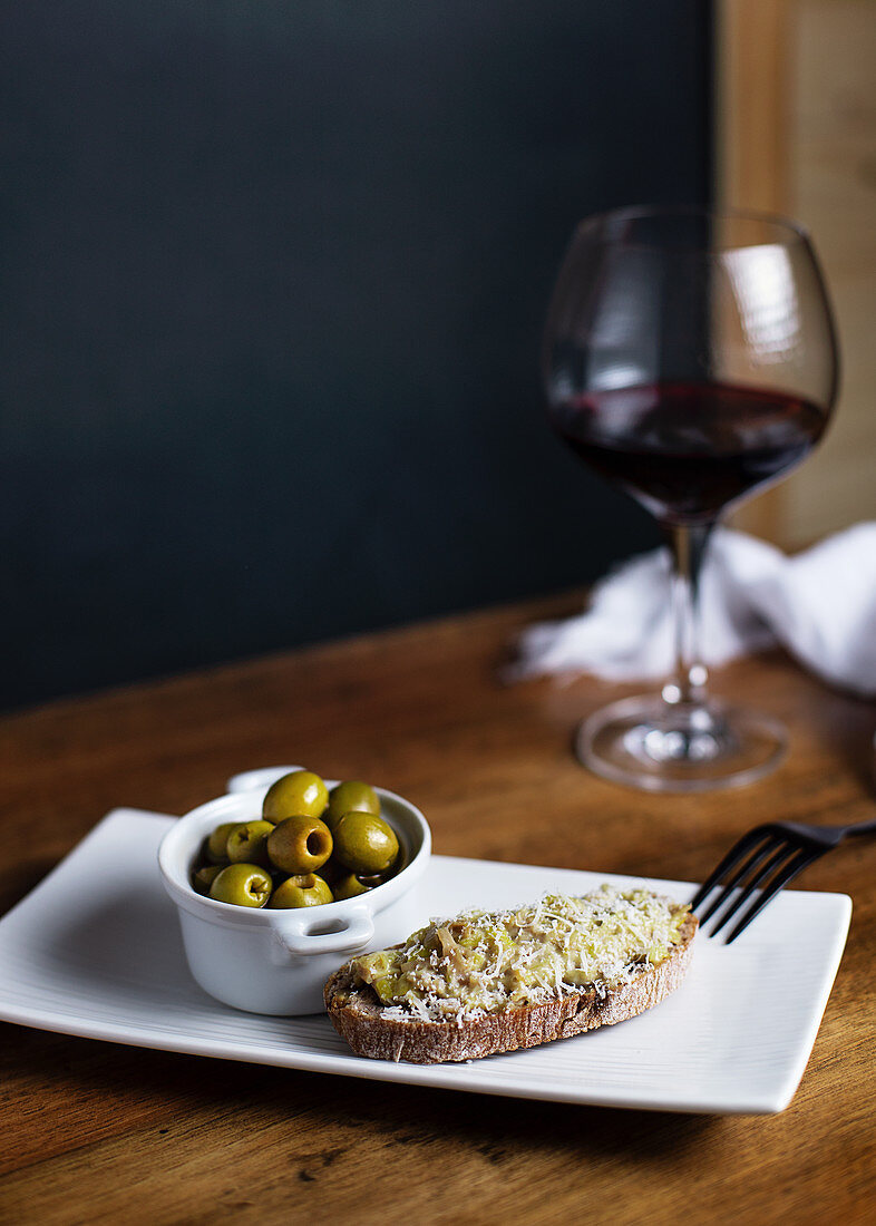 Bowl with pickled olives and bruschetta on plate placed on table with glass of red wine