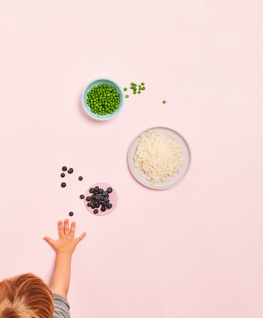 A child's hand reaching for rice, peas and blueberries