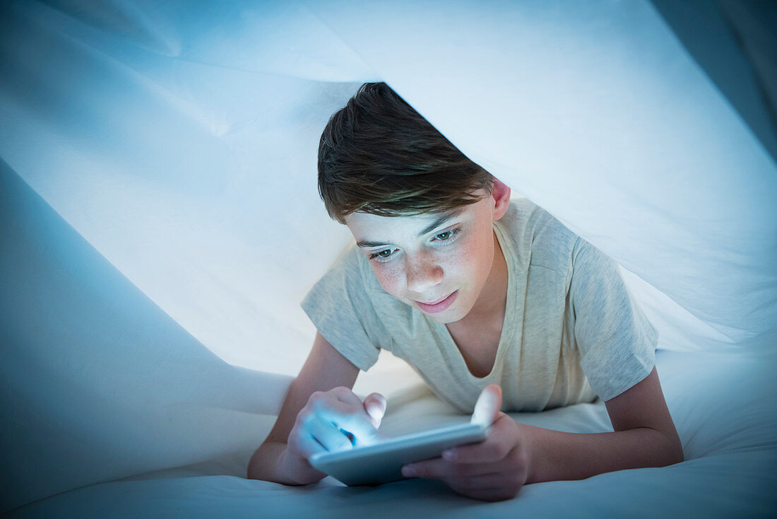 Boy using digital tablet under sheet