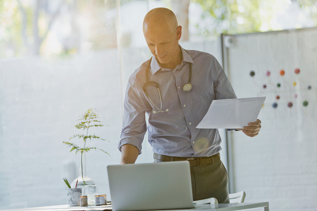 Male doctor with paperwork standing at laptop