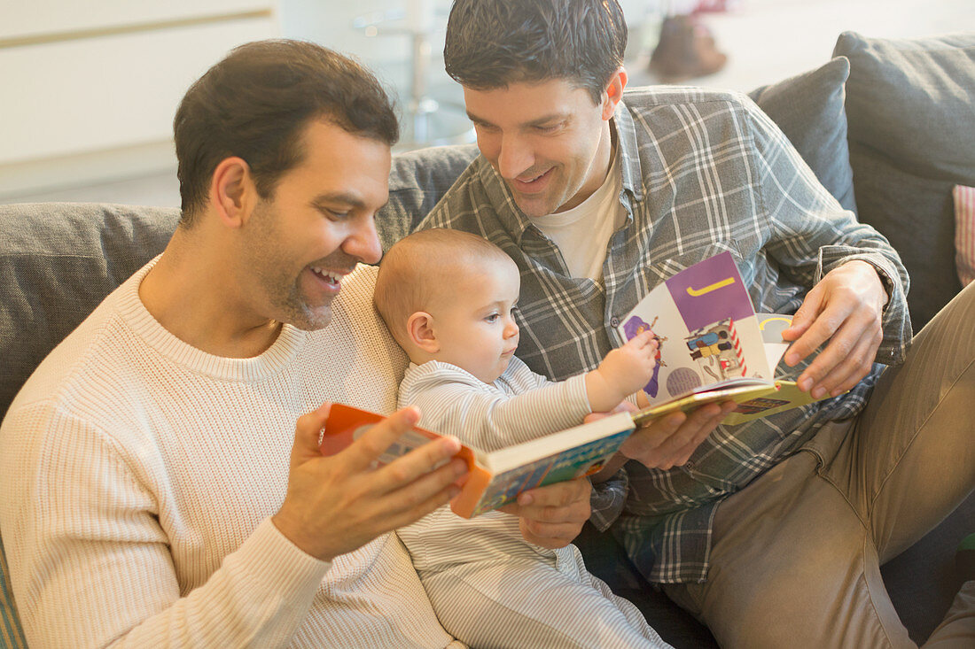 Male gay parents reading book to baby son on sofa