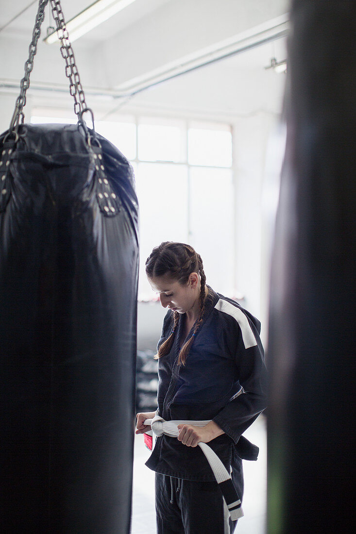 Young woman tying jiu-jitsu belt next to punching bag