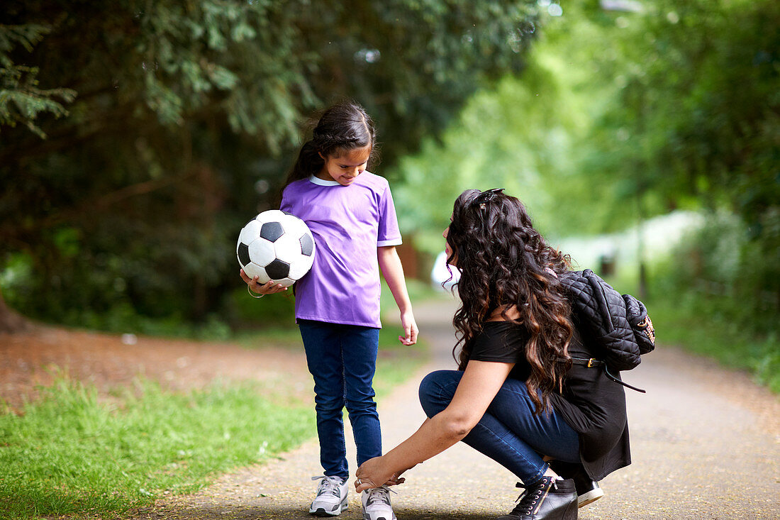 Mother tying shoelace of daughter holding ball