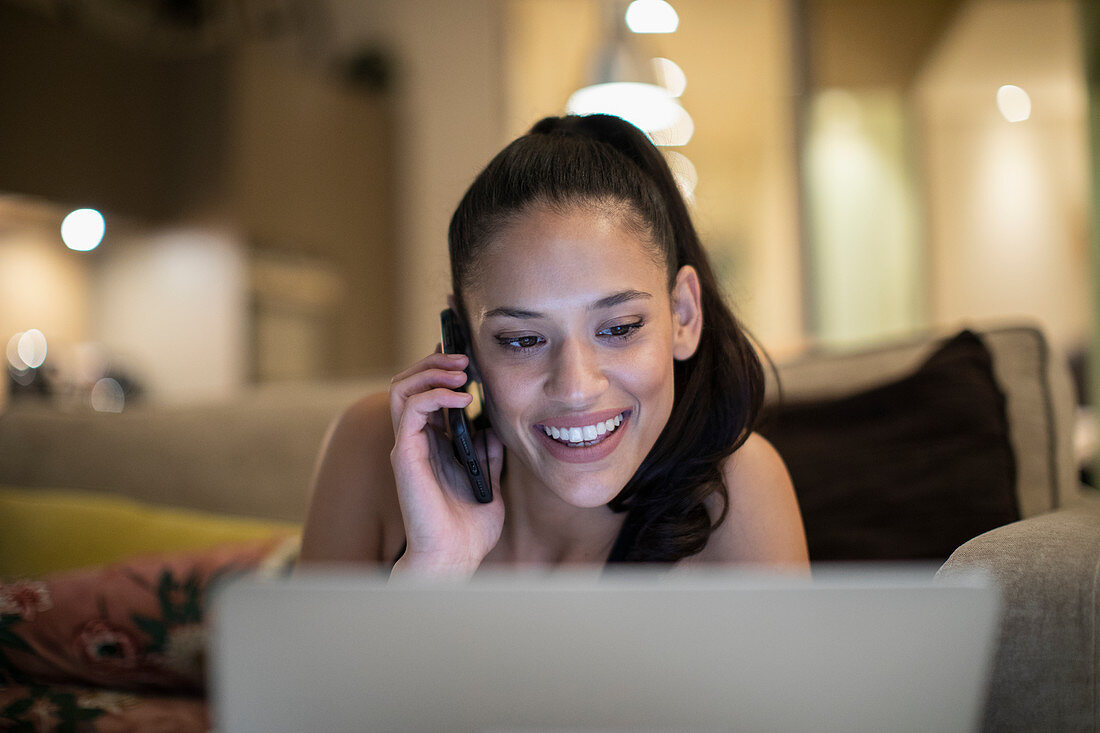 Smiling woman talking on smart phone at laptop on sofa