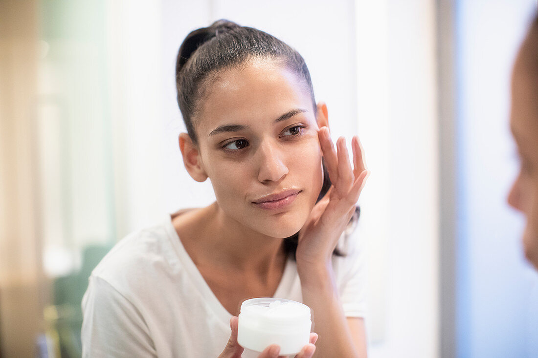Woman applying moisturizer to face in bathroom mirror