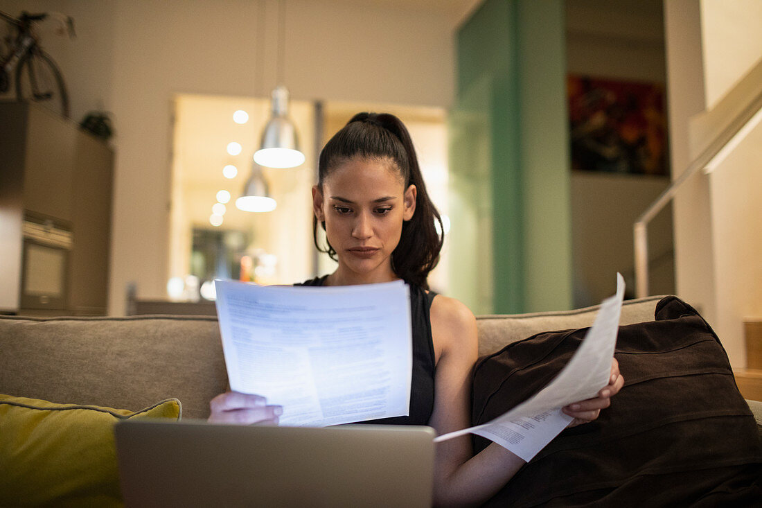 Woman reading paperwork, working at laptop on sofa