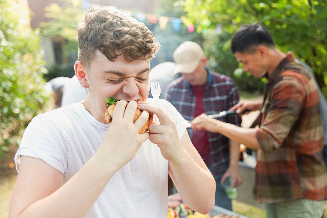 Hungry teenage boy eating hamburger at backyard barbecue