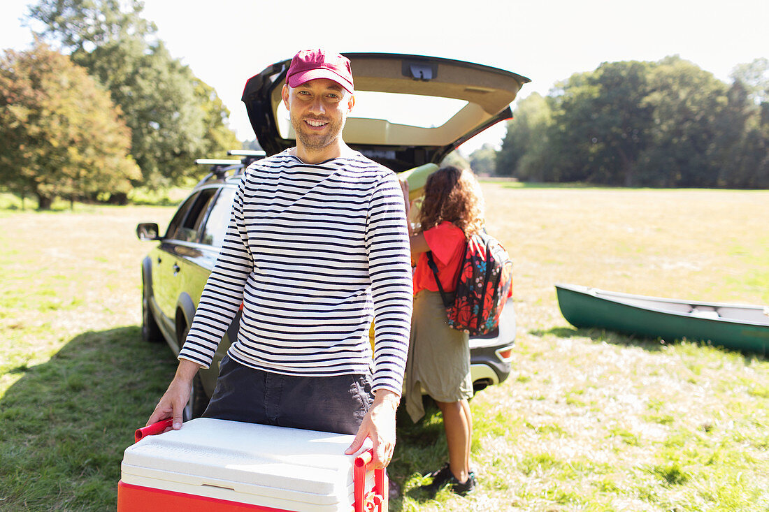 Portrait man carrying camping cooler, unloading car in field