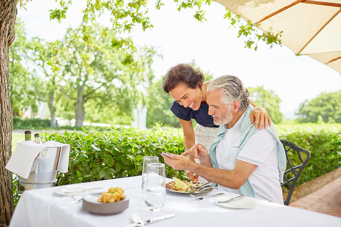 Mature couple dining, using smart phone at patio table