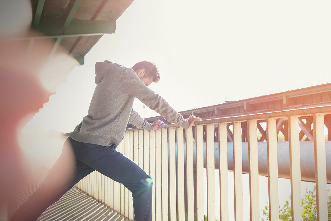 Young male runner stretching leg at sunny urban railing