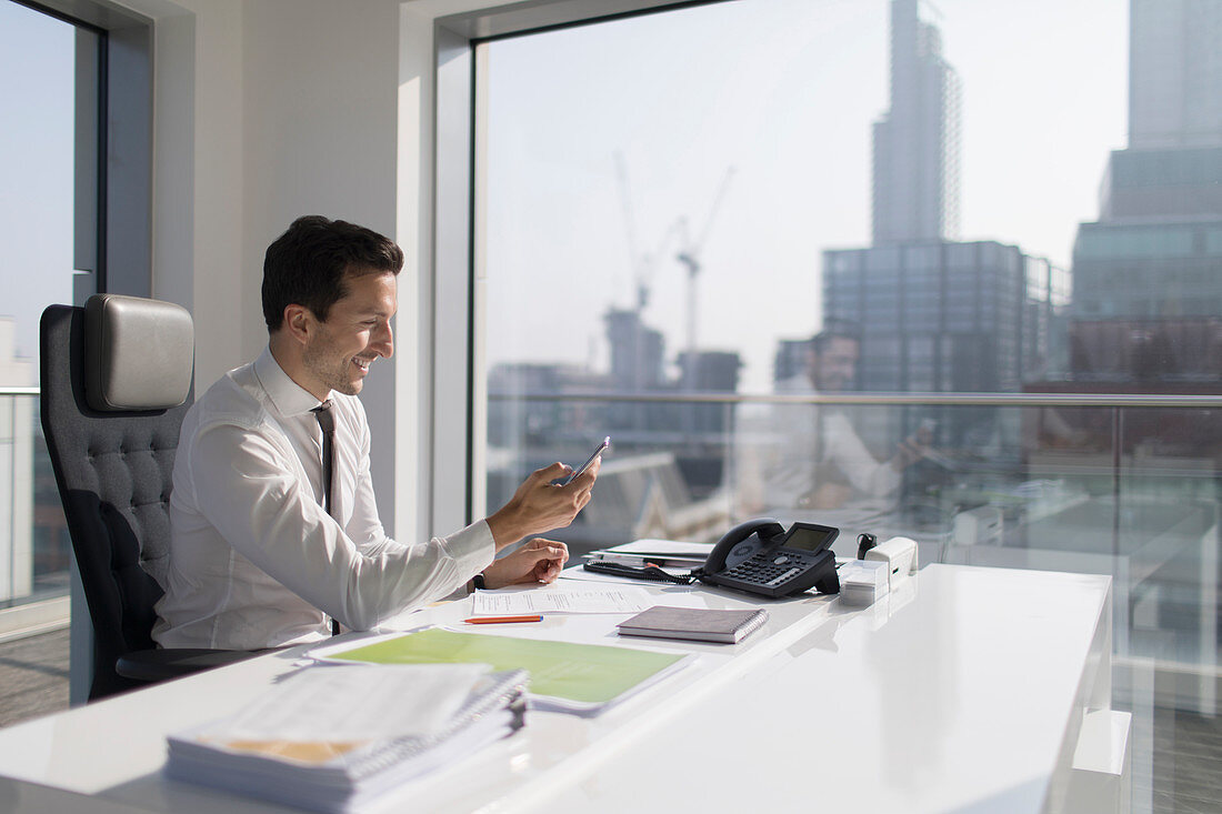 Businessman using smart phone in sunny, urban office