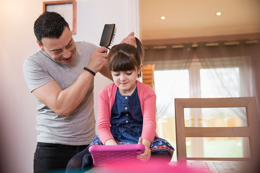 Father brushing hair of daughter using digital tablet