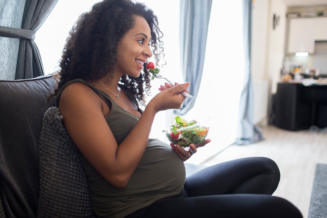 Smiling pregnant woman eating salad on sofa