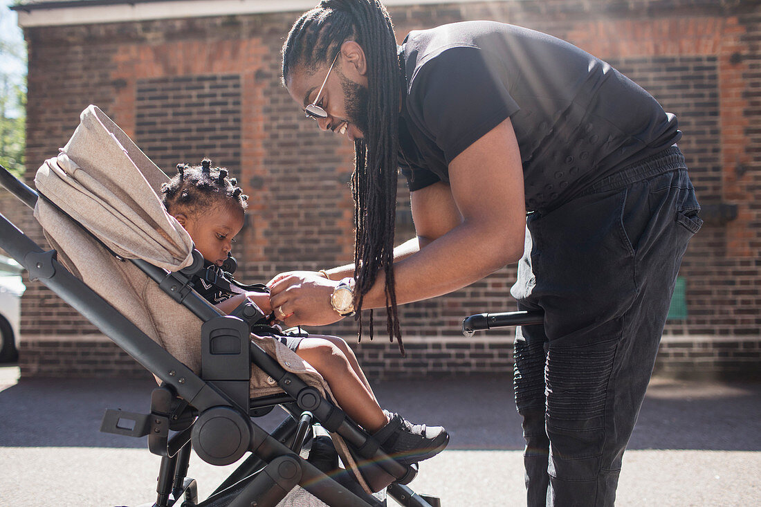 Father fastening toddler son in stroller on sidewalk