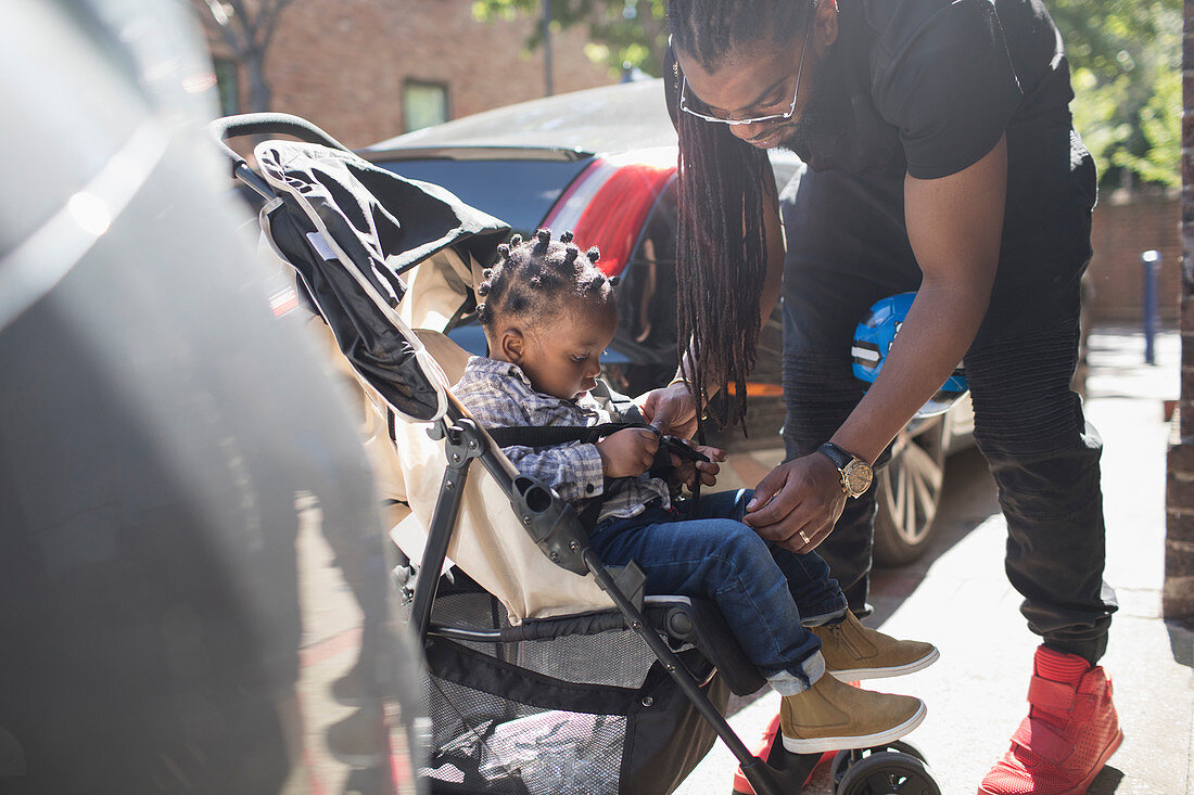 Father fastening toddler son in stroller on sidewalk